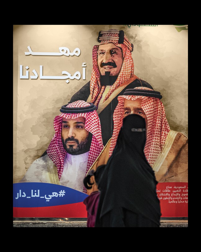 woman in niqab and abaya looks at camera while walking with a child past a larger-than-life-size poster of the royal family