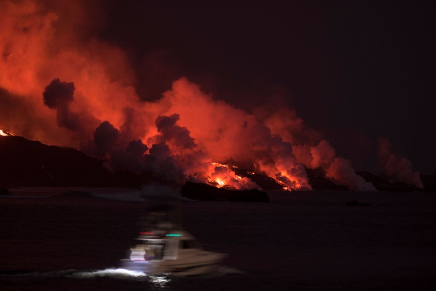 Nighttime view of lava flowing into the ocean, glowing orange, and sending up plumes of steam.