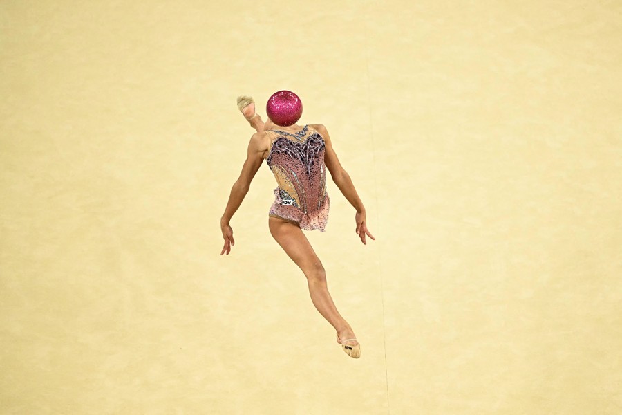 A gymnast jumps while leaning back, with a ball sitting where one would expect her head to be.