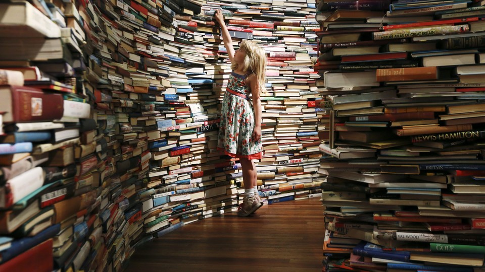 A young girl reaches for a book, surrounded by hundreds of other books.