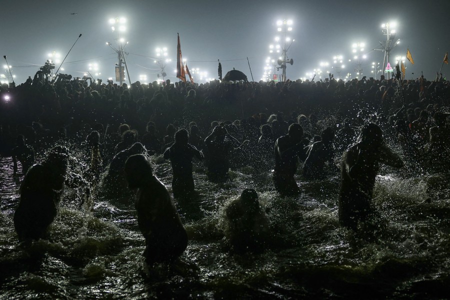Many men splash and dunk themselves in shallow water, with a large crowd and lamp posts visible in the background.