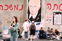 Photograph of a wall with an illustration of Yitzhak Rabin, Hebrew writing, and mourning protesters sitting in front of it