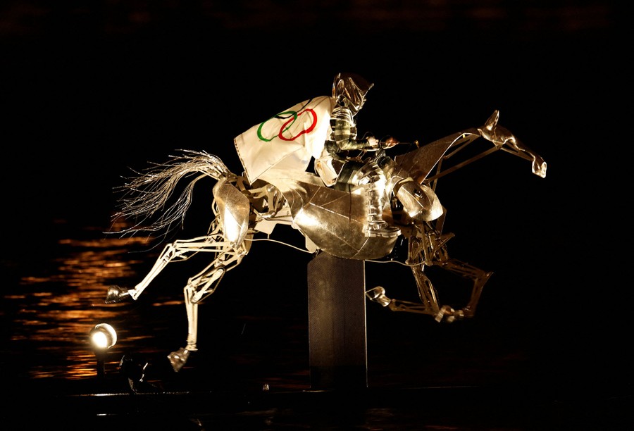 An armored woman on a robotic horse wears the Olympic Flag as a cape while crossing a river at night.