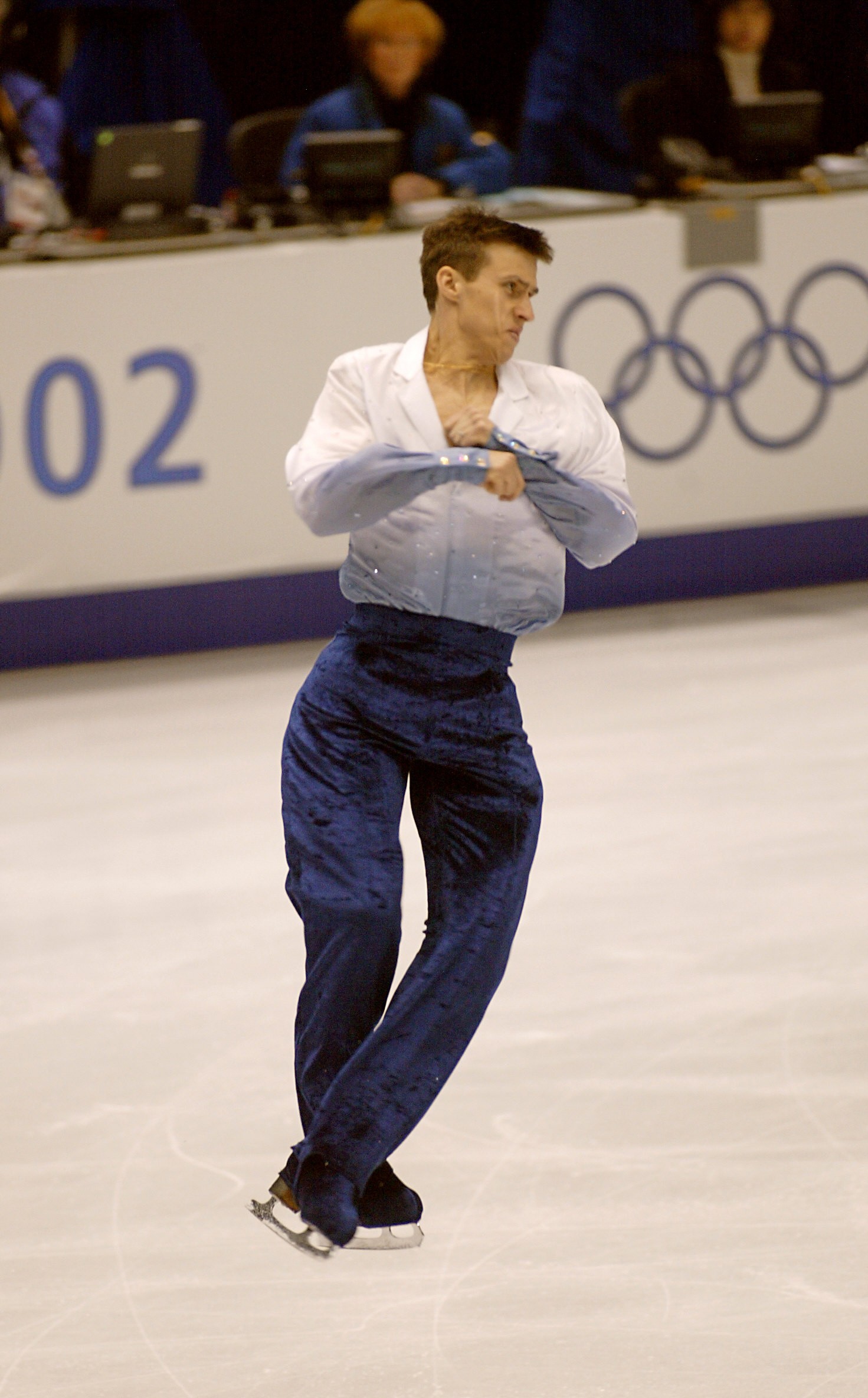 photo of man in white shirt and blue pants mid-spin in ice rink with Olympic logo in background