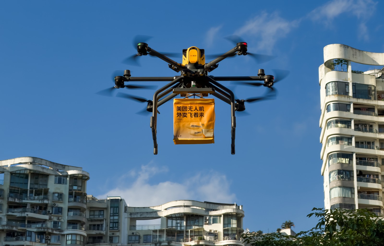 A six-rotor drone carries a takeaway delivery bag over residential buildings.