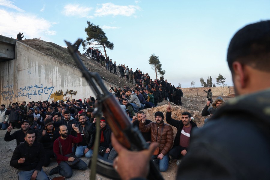 Dozens of men in civilian clothes sit on the ground beside a road bridge, under the guard of an armed man in the foreground.