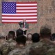 Donald Trump speaks to troops at al Asad Air Base with an American flag behind him and Melania Trump next to him.