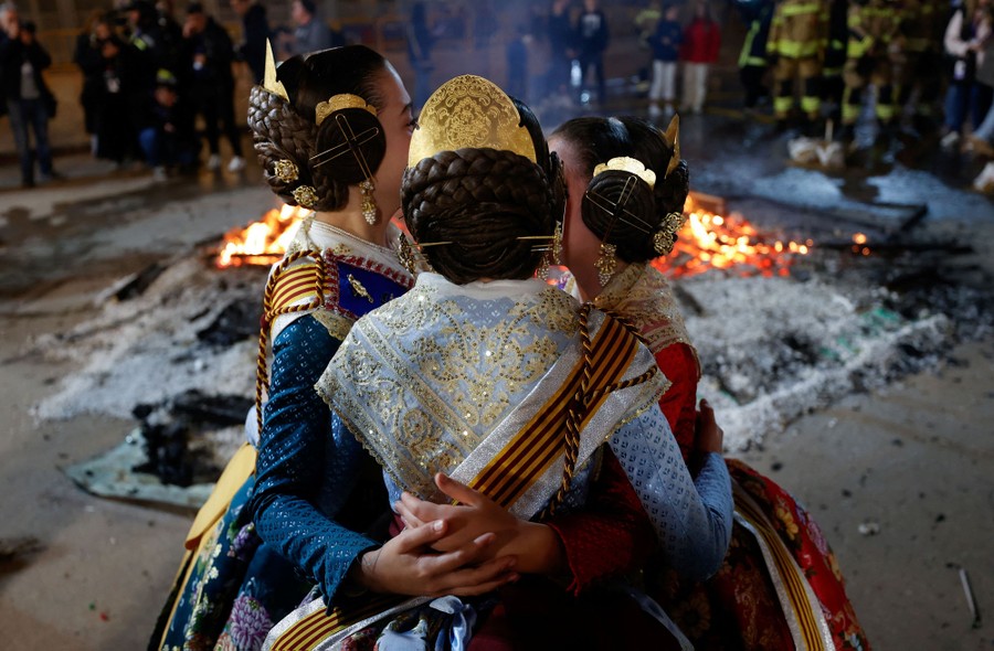 Three girls wearing traditional clothing embrace beside a pile of ash and glowing embers.