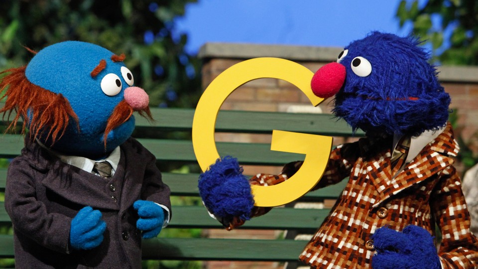 Grover holds up a yellow "G" as Fat Blue looks at him. They sit on a park bench. 