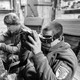 In a black-and-white photo, two men sit at a table operating drones.