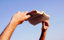 Two hands hold a paperback book open against the sky.