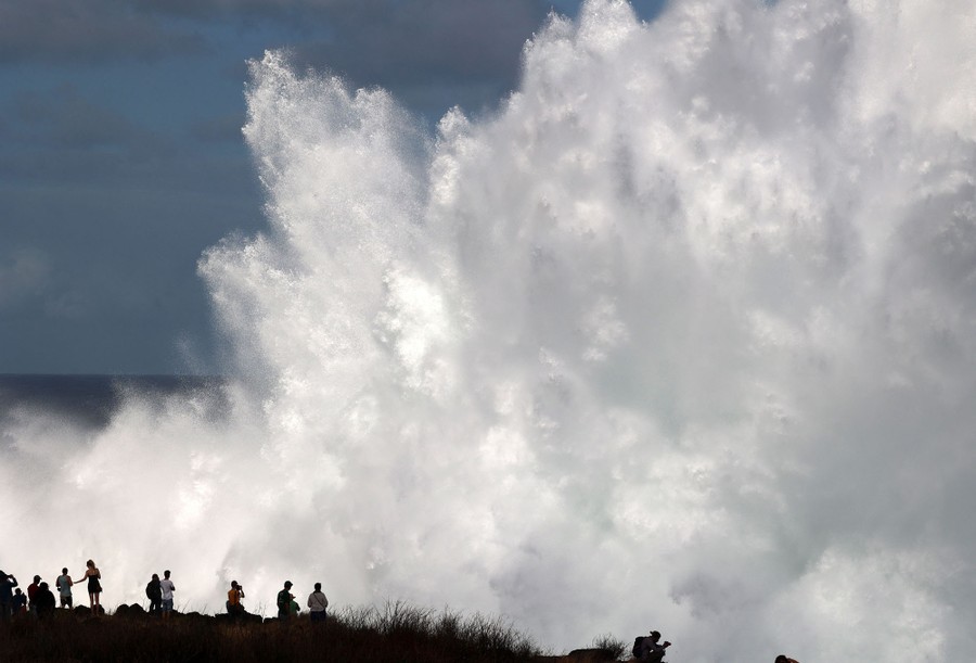 People stand near a shore, watching huge plumes of water thrown high by crashing waves.