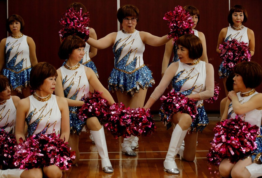 About a dozen older women dressed in showy cheerleader costumes pose together in a gymnasium.