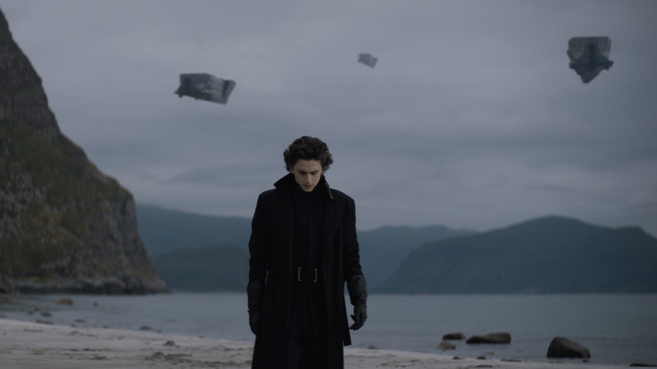 Timothée Chalamet standing on a beach, dressed in a thick black coat, pieces of something blowing in the wind against a cloudy sky