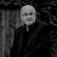A black-and-white photograph of Salman Rushdie sitting on a park bench.