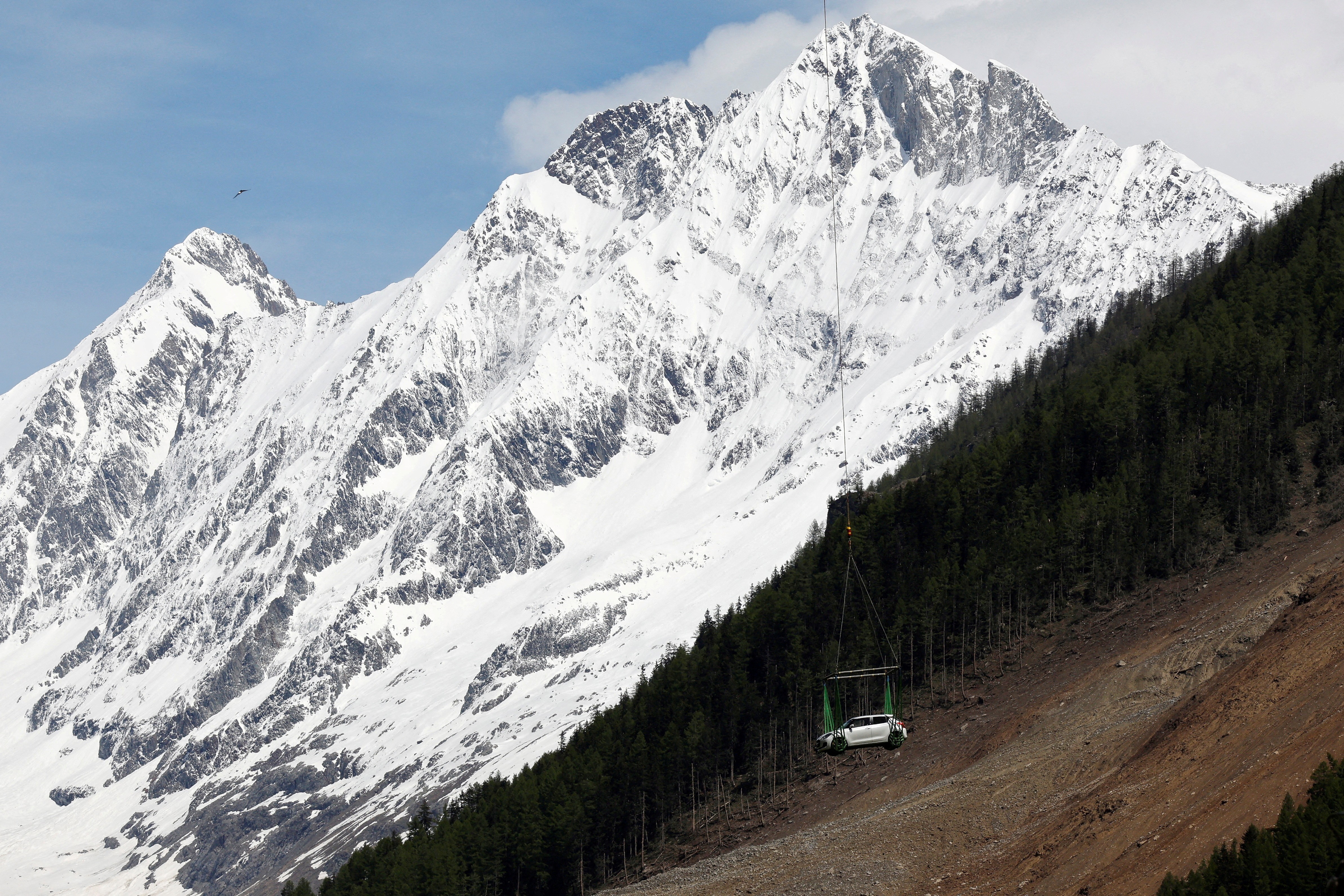 A car hangs from a sling beneath a helicopter (unseen), in front of a large snow-covered mountain.