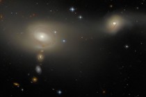 A pair of interacting galaxies, one smaller than the other. They appear as a broad, soft glow in which individual stars aren't visible. A number of bright stars and smaller, background galaxies are also featured—three such galaxies lie in a vertical line below the right-hand galaxy of the pair.