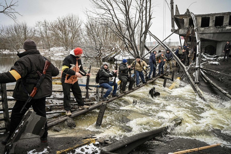 A line of about a dozen people can be seen sitting on the wreckage of a destroyed bridge across a small river, each passing a rifle to the next.
