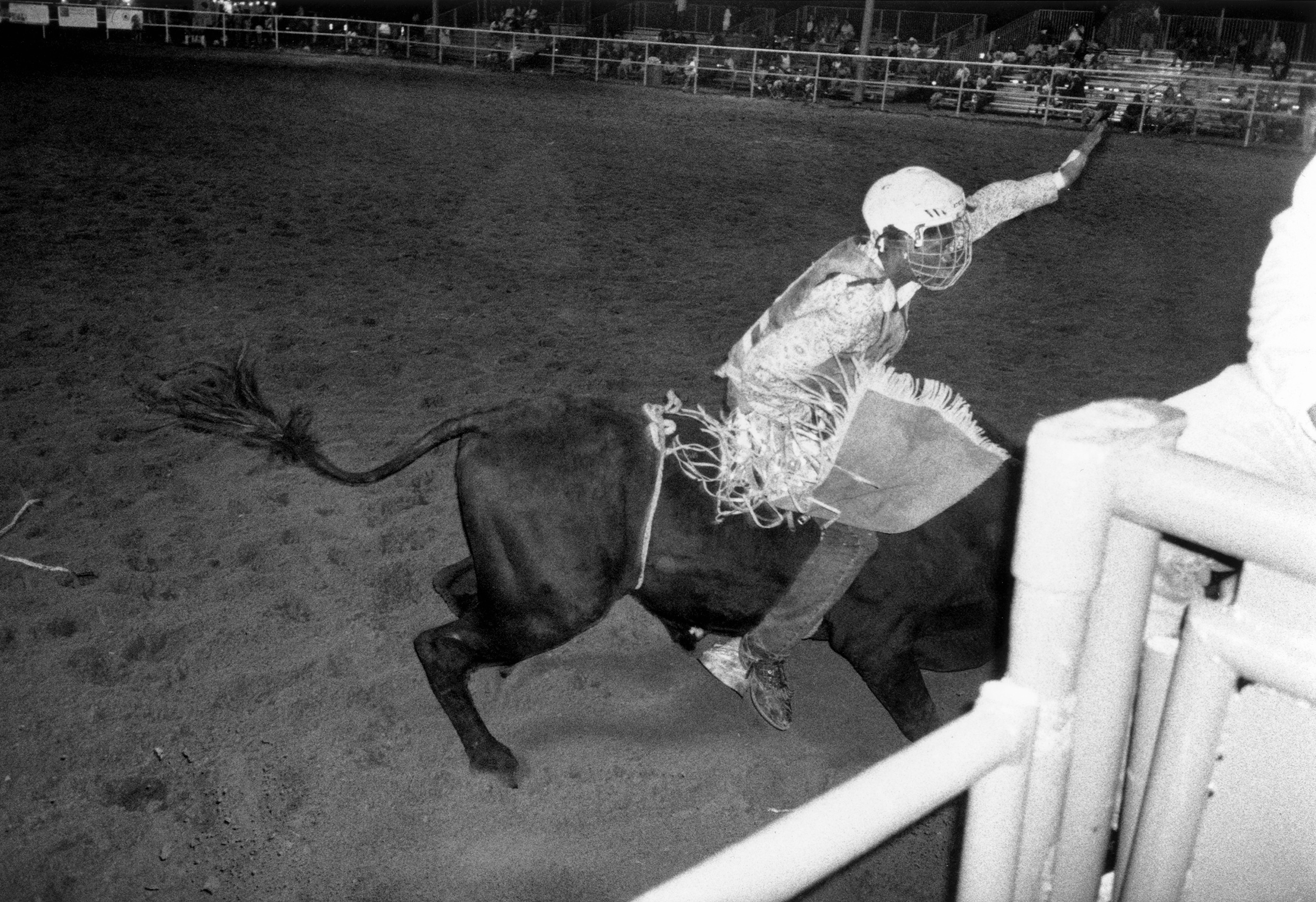 black-and-white photo of bull rider in helmet and fringed chaps riding in arena 