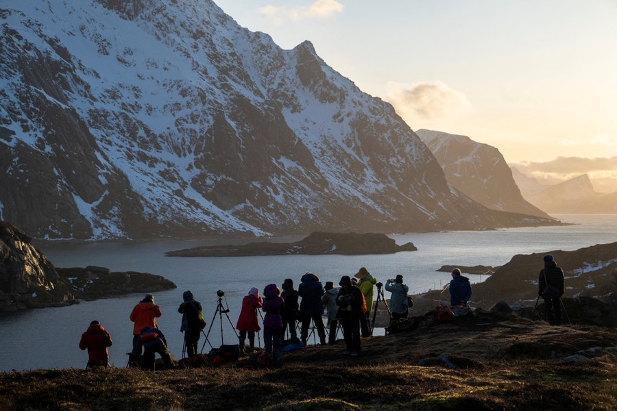 Tourists line up to photograph a fjord and steep snow-covered mountains.