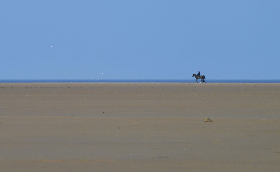 Two people and a horse walk on a wide, empty stretch of sandy beach.