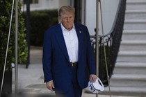 Donald Trump walking, looking displeased, carrying in one hand a hat that reads "USA"