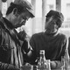 A black and white image of two men in a bar smoking. One man offers the other a light.
