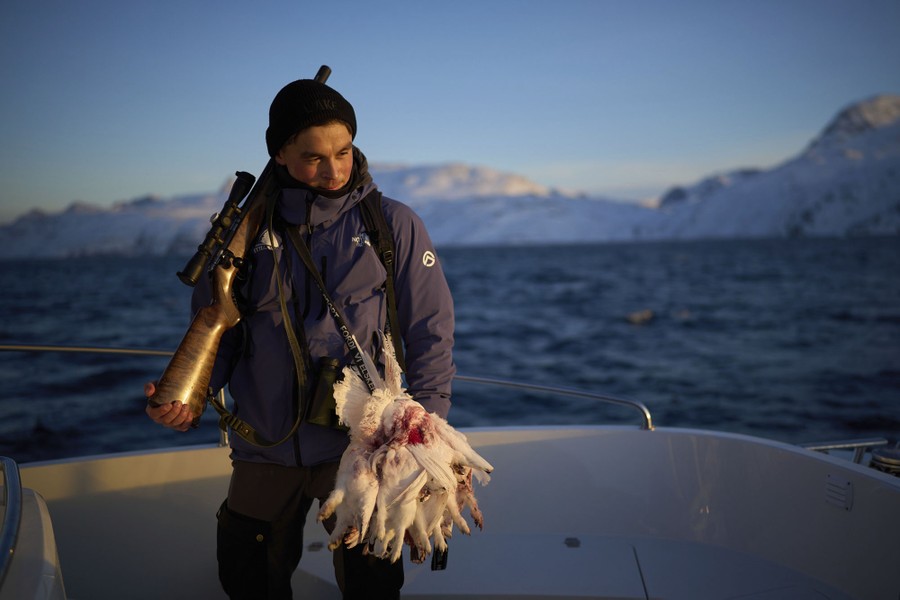 A hunter holds a rifle over their shoulder and a clutch of hunted ptarmigan in one hand, standing on a boat.