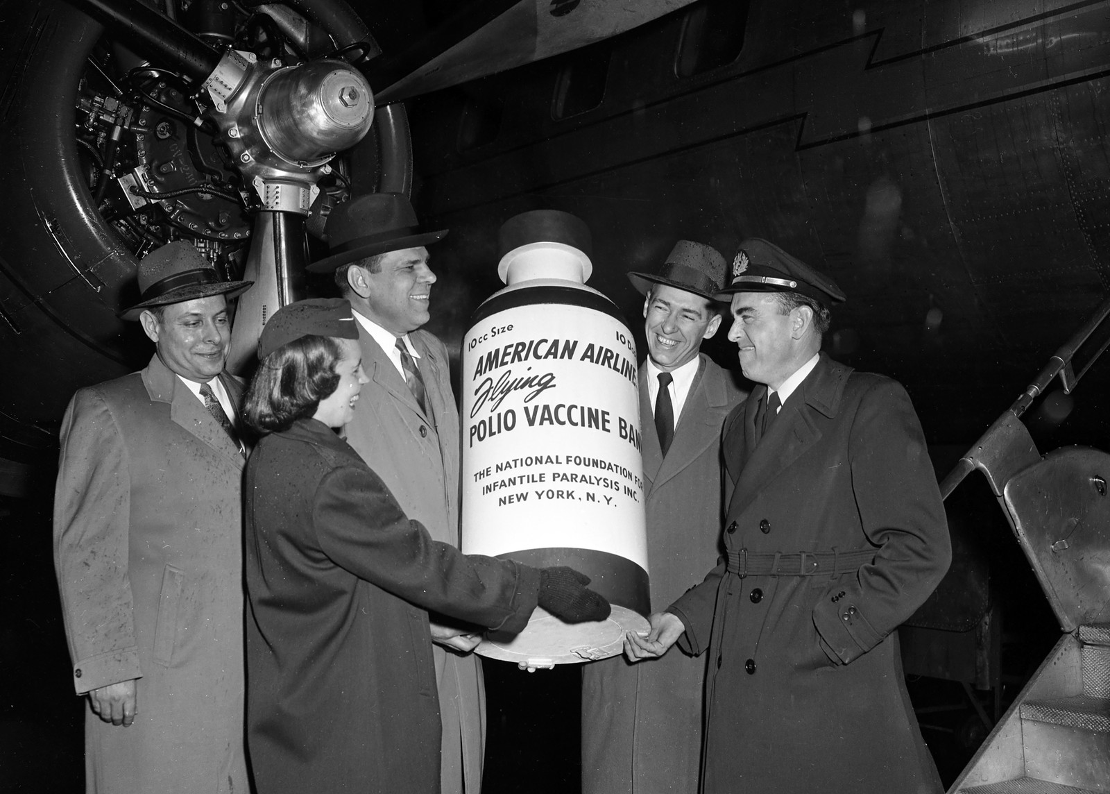 Five people pose outside of a propeller aircraft, holding up an oversized vaccine bottle promoting American Airlines and the polio vaccine.