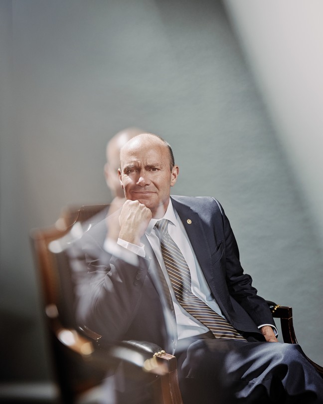 A color portrait of Mike Lee in suit and tie, sitting in chair with hand to chin, with double exposure of image