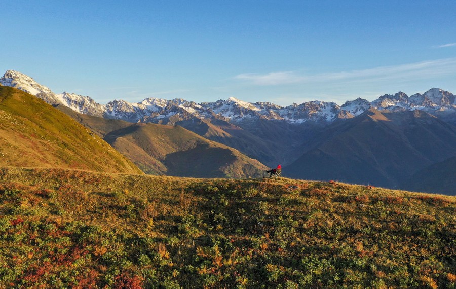 A person sits in a chair along a ridge among high mountains.