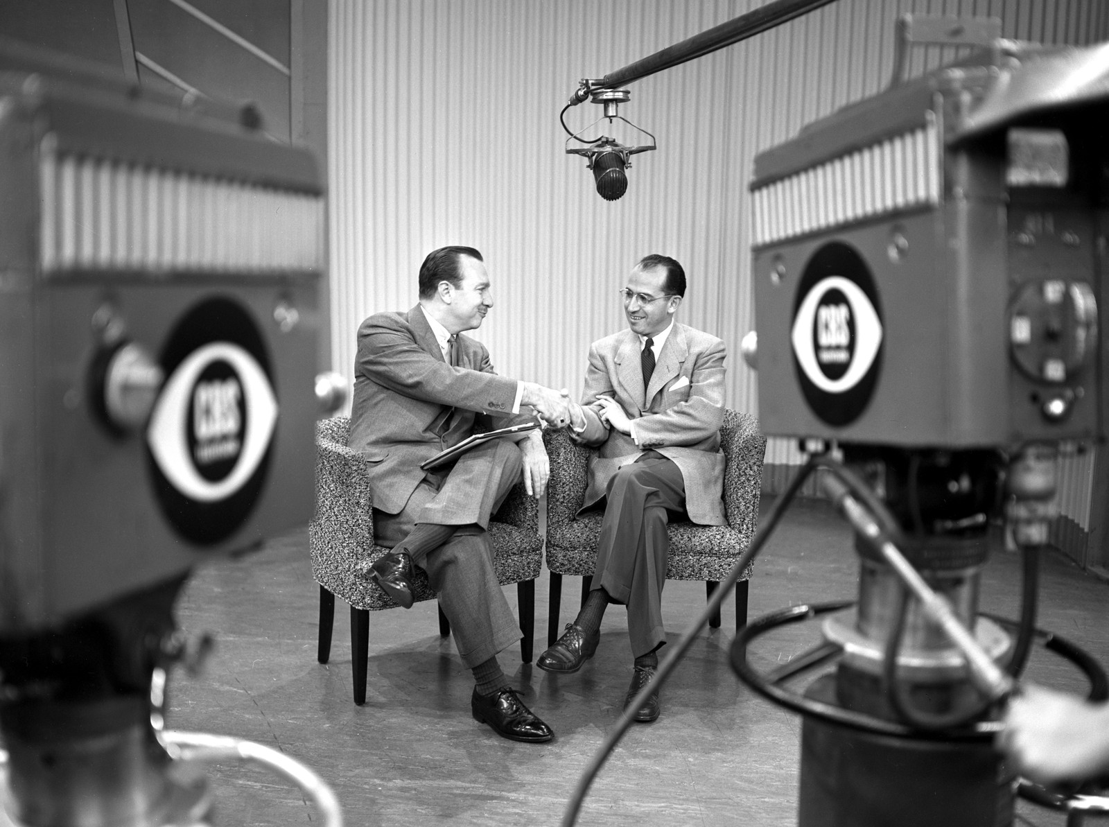 Two men sit in a comfortable chairs in a studio, shaking hands in front of two large television cameras.