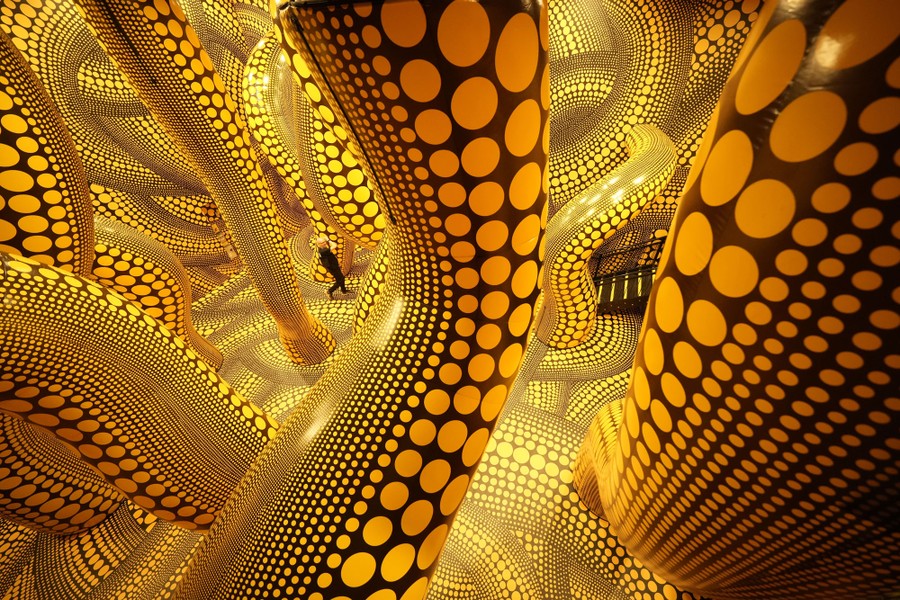 A person walks through an art installation—a large room filled with long, inflatable, tentacle-like tubes. Everything is painted with yellow dots on a black background.