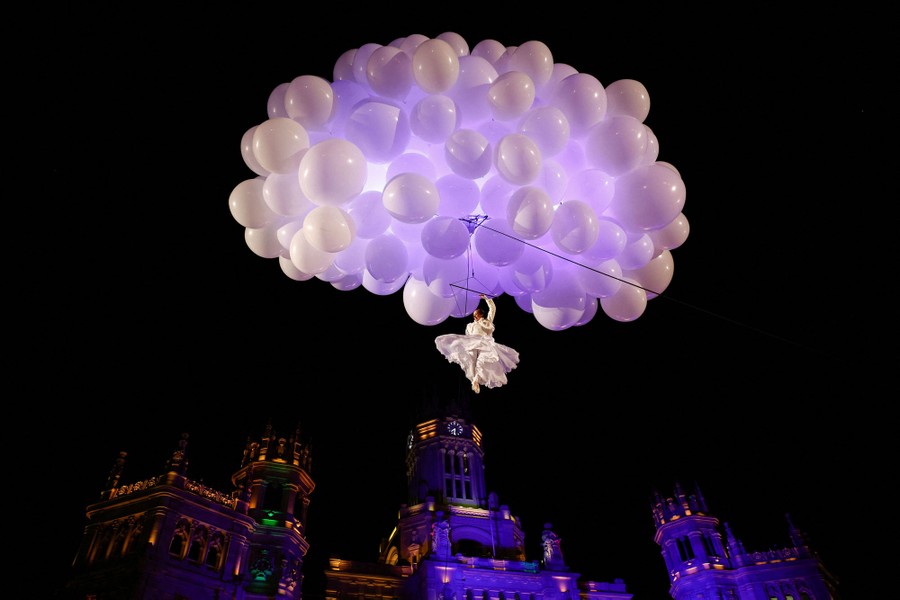 A performer hangs from a wire beneath a cloud-shaped cluster of white balloons.