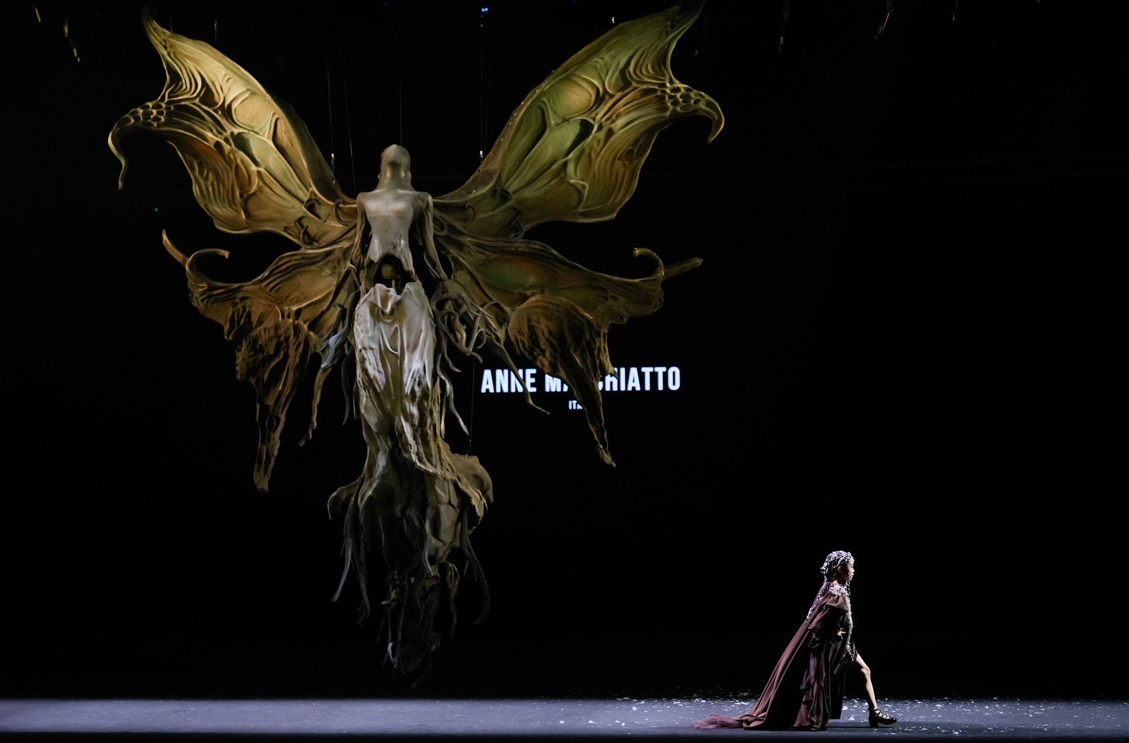 A young model walks on a runway during a fashion show beneath a sculpture of a winged fairy-like being.