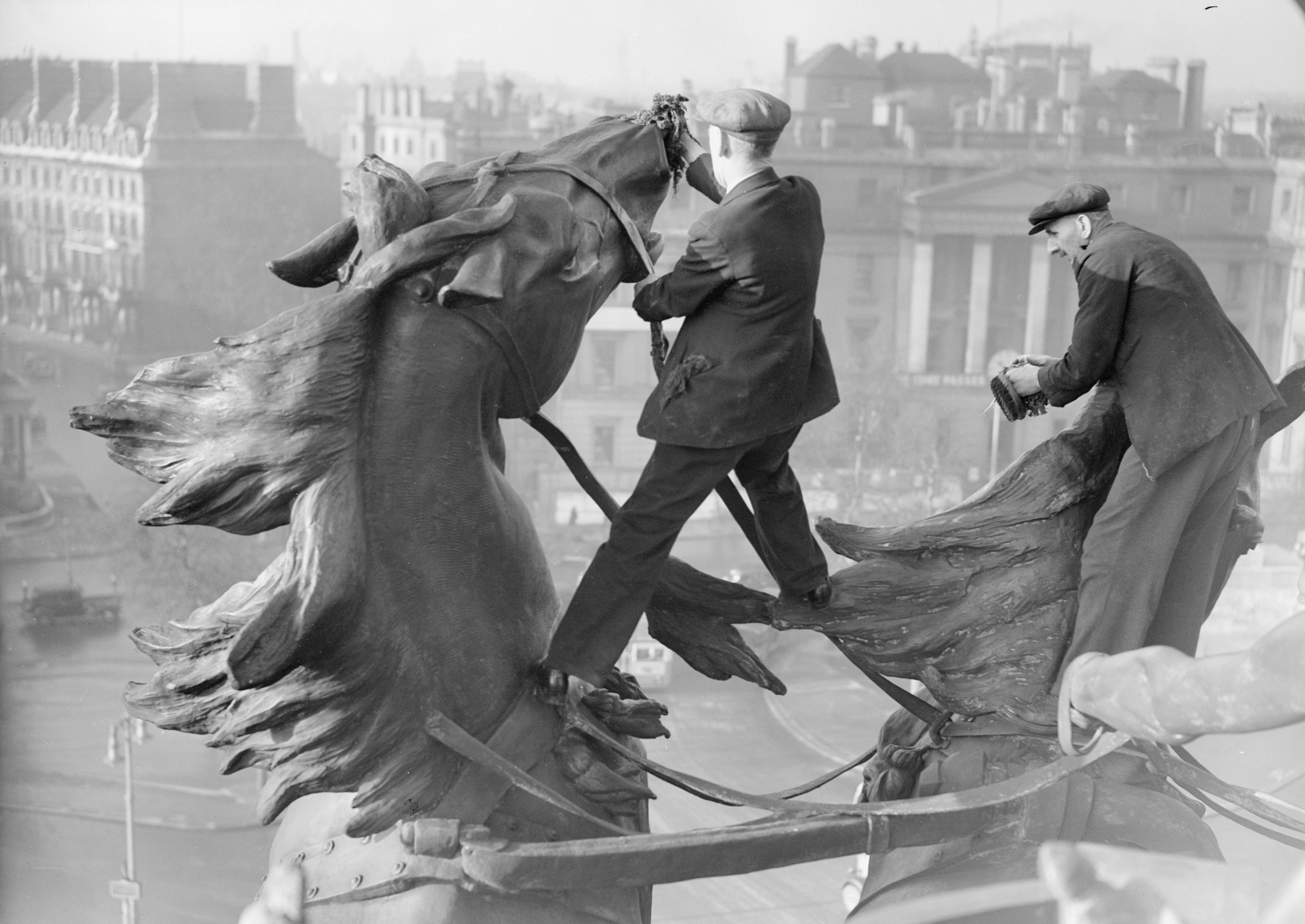 Two worker clean a large statue of horses atop a tall arch in 1939.