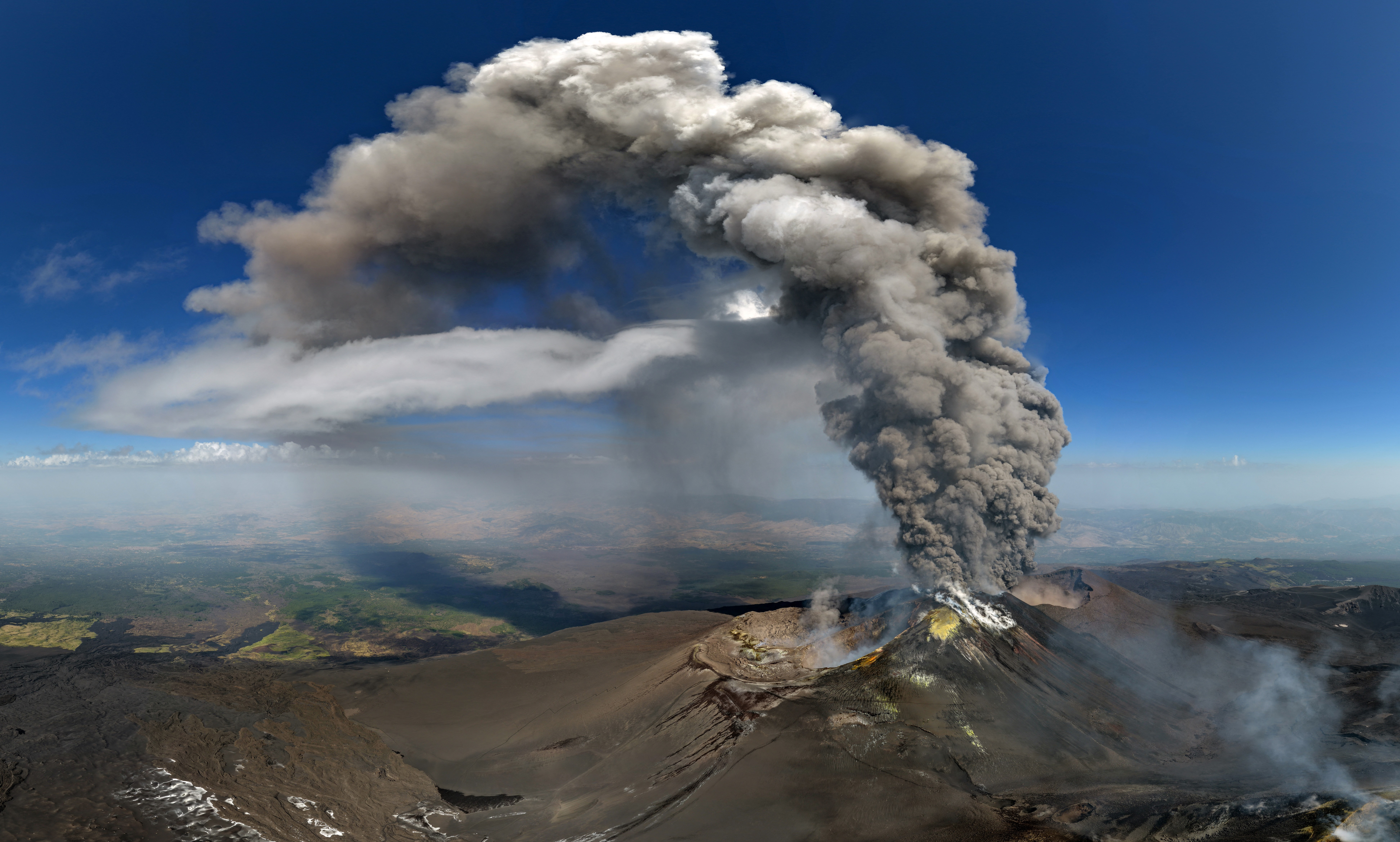 A plume of ash rises from Mount Etna, arcing into the distance as wind carries it away.
