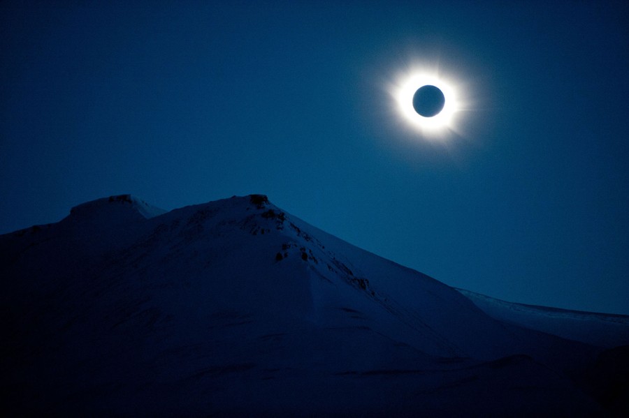The bright corona of the sun radiates out from the black circle made by the moon as it passes in front of the sun, seen above a snowy mountain.