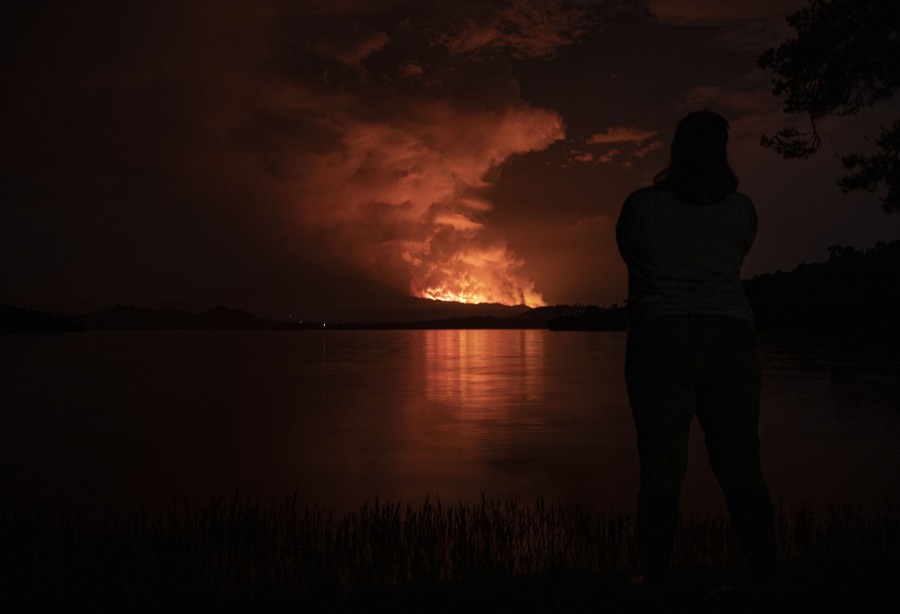 A person stands near the shore of a lake at night, observing a glow and rising smoke from a lava flow in the distance.