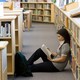 Asian American student sitting on the floor of a library reading a book, leaning back against the shelves