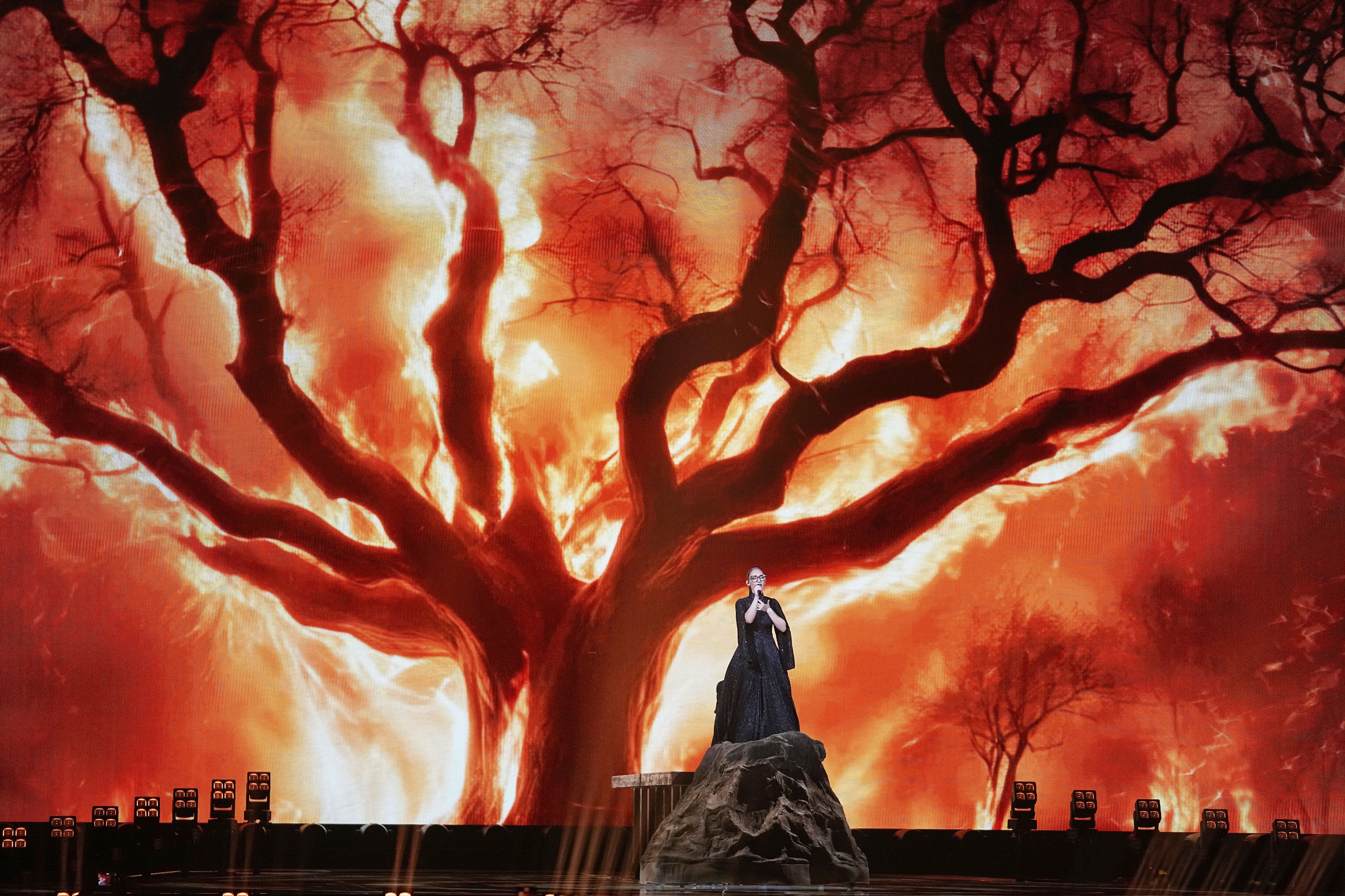 A singer performs on a vast stage in front of a huge video image of a huge burning tree.