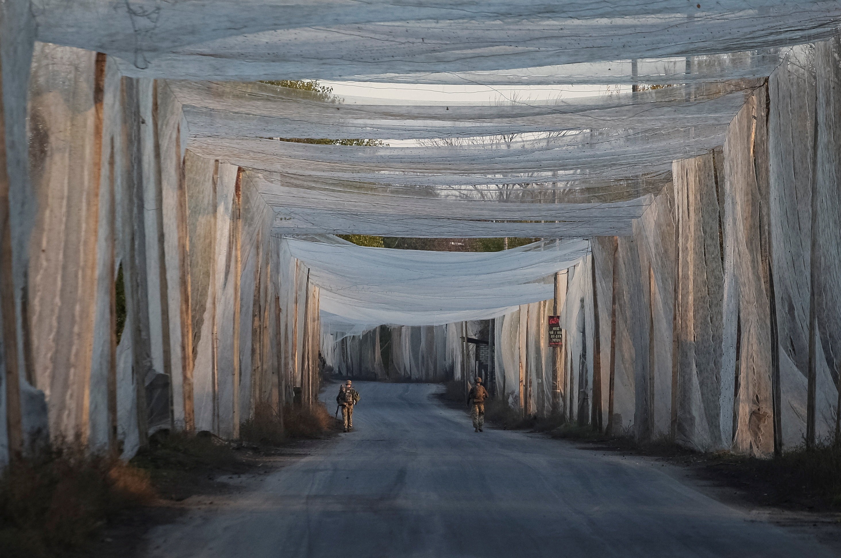 Two Ukrainian servicemen walk along a road covered with anti-drone netting.