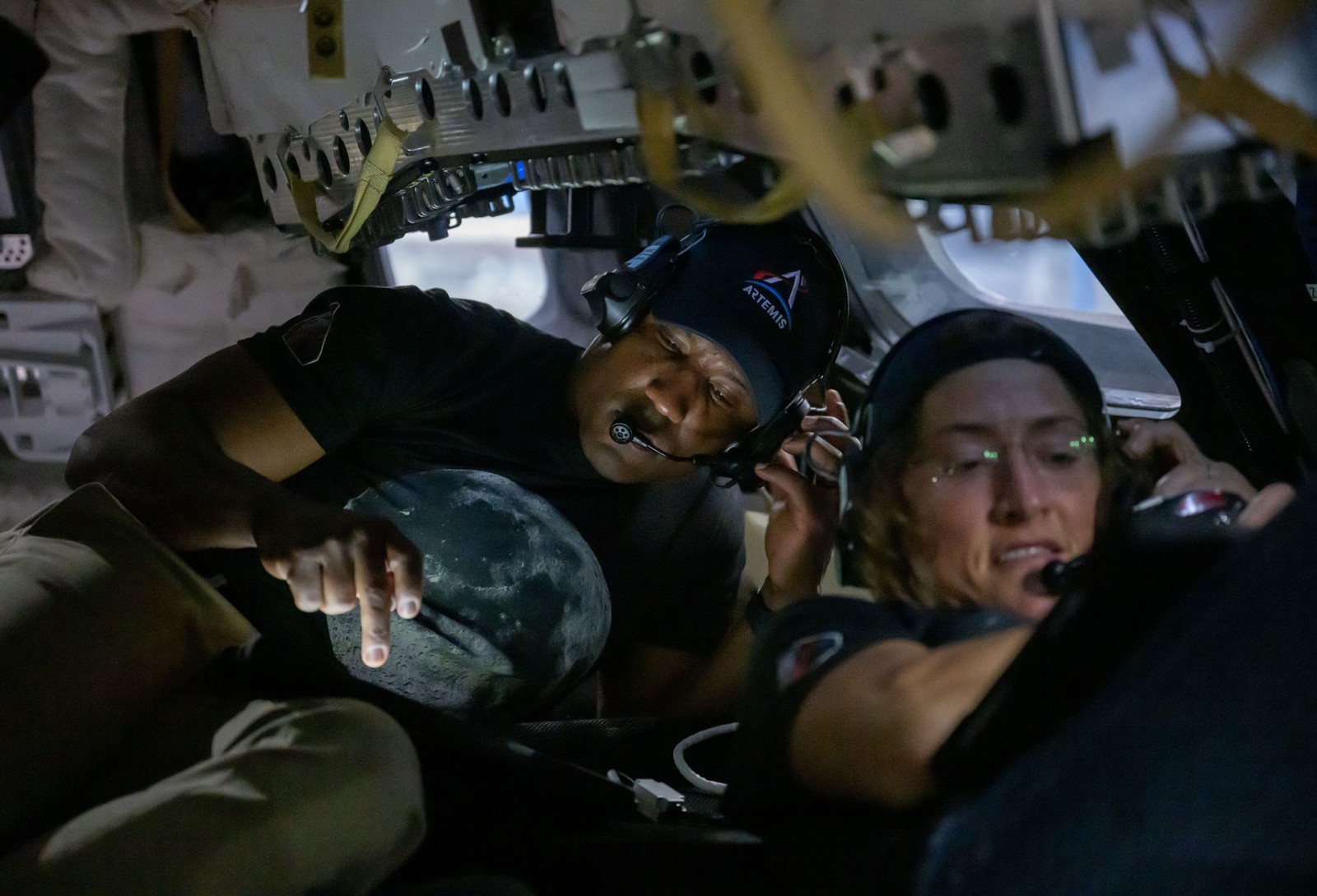 Two astronauts train inside a capsule mockup, communicating with headsets.