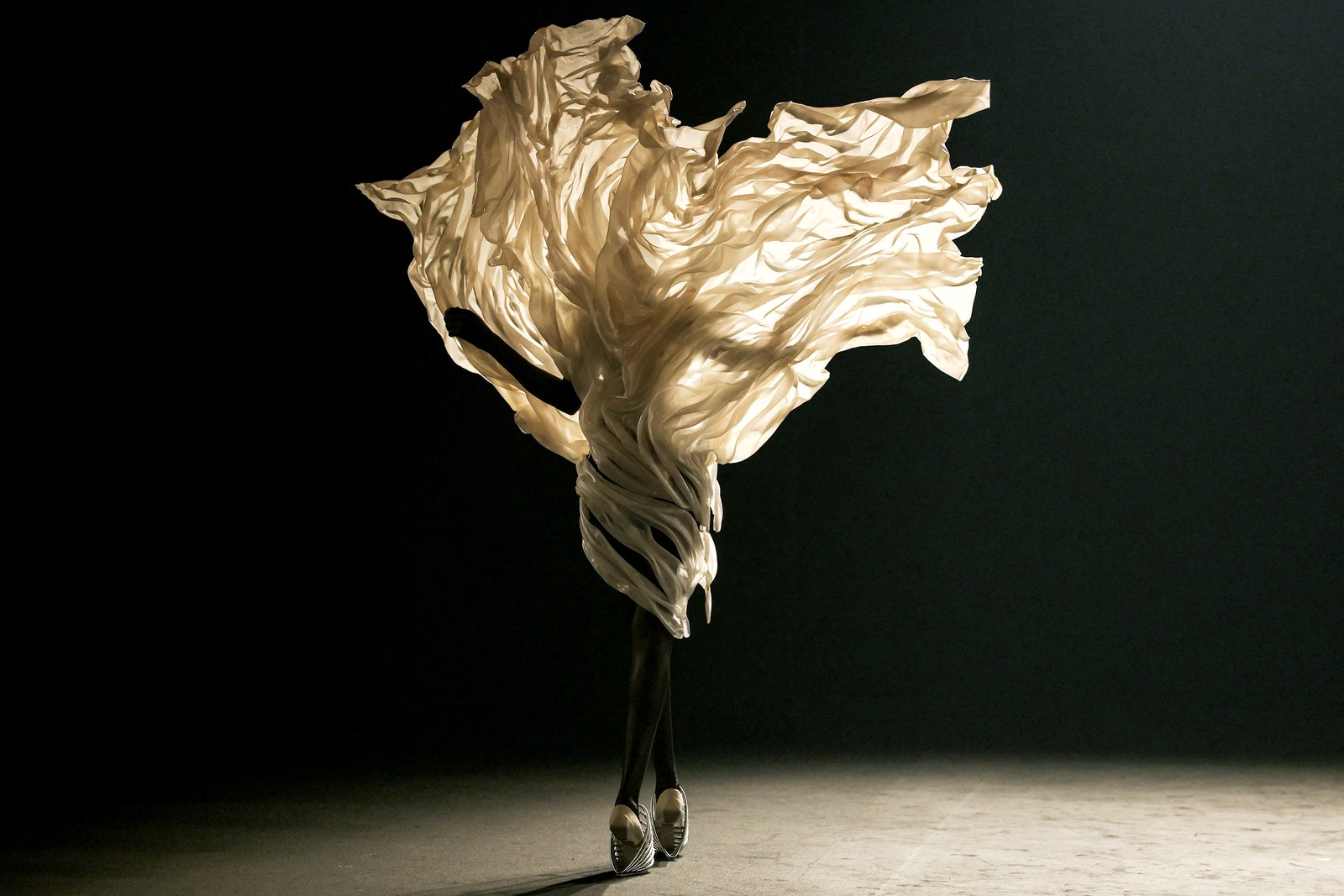 A model presents a garment that appears to be a large piece of fabric being blown upward by a strong gust of wind.