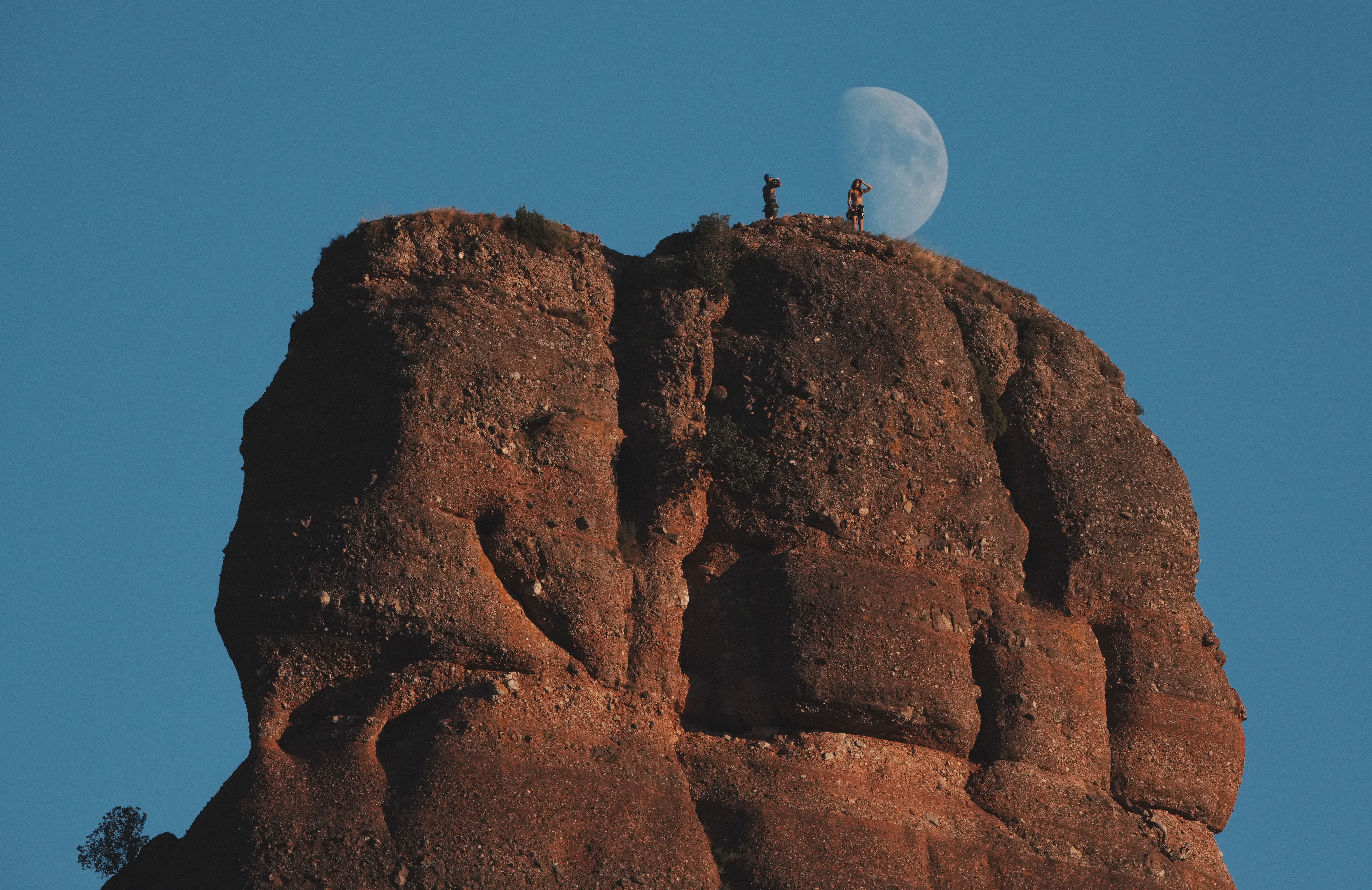 Two climbers stand atop, a monolithic rock formation, with the moon visible in the blue sky behind them.