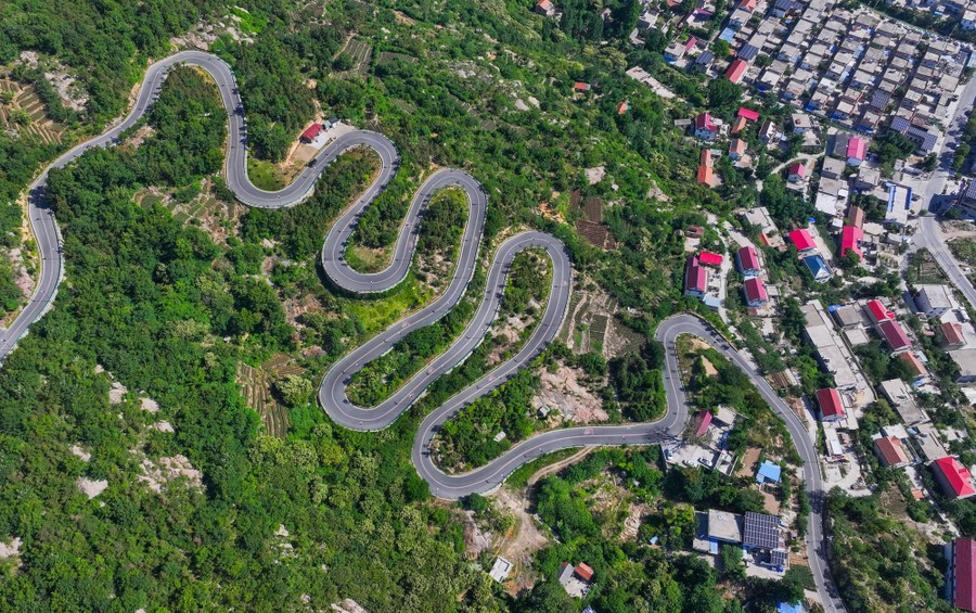 A winding road climbs a hill above a village, seen from above.