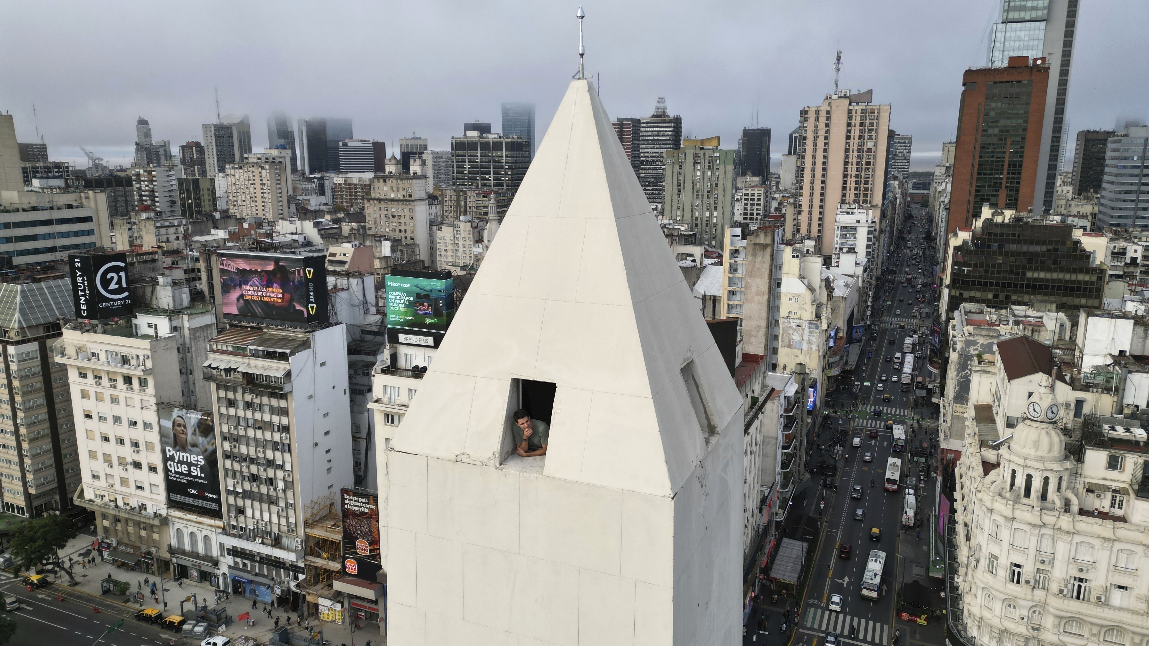 A person peers out of a window atop a tall obelisk in the middle of a city.