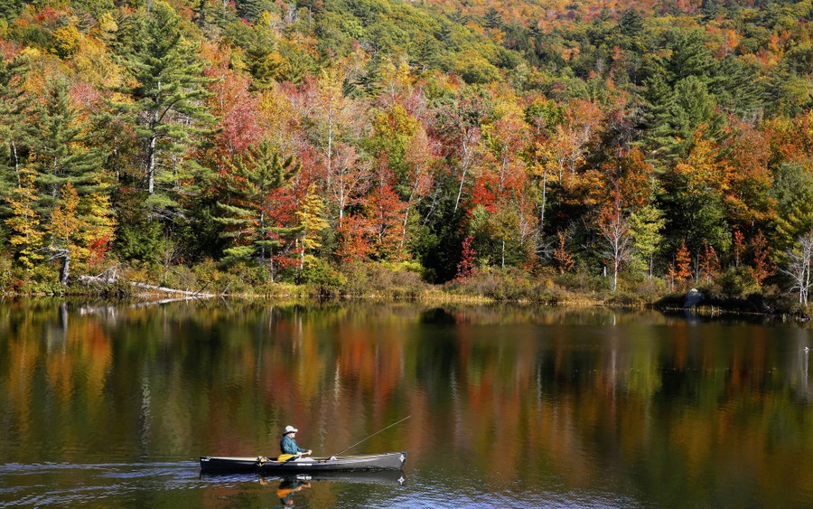 A fly fisherman paddles on a pond as fall foliage begins to show color.