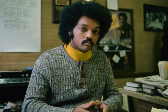 A color photograph of Jesse Jackson in his office wearing a yellow turtleneck