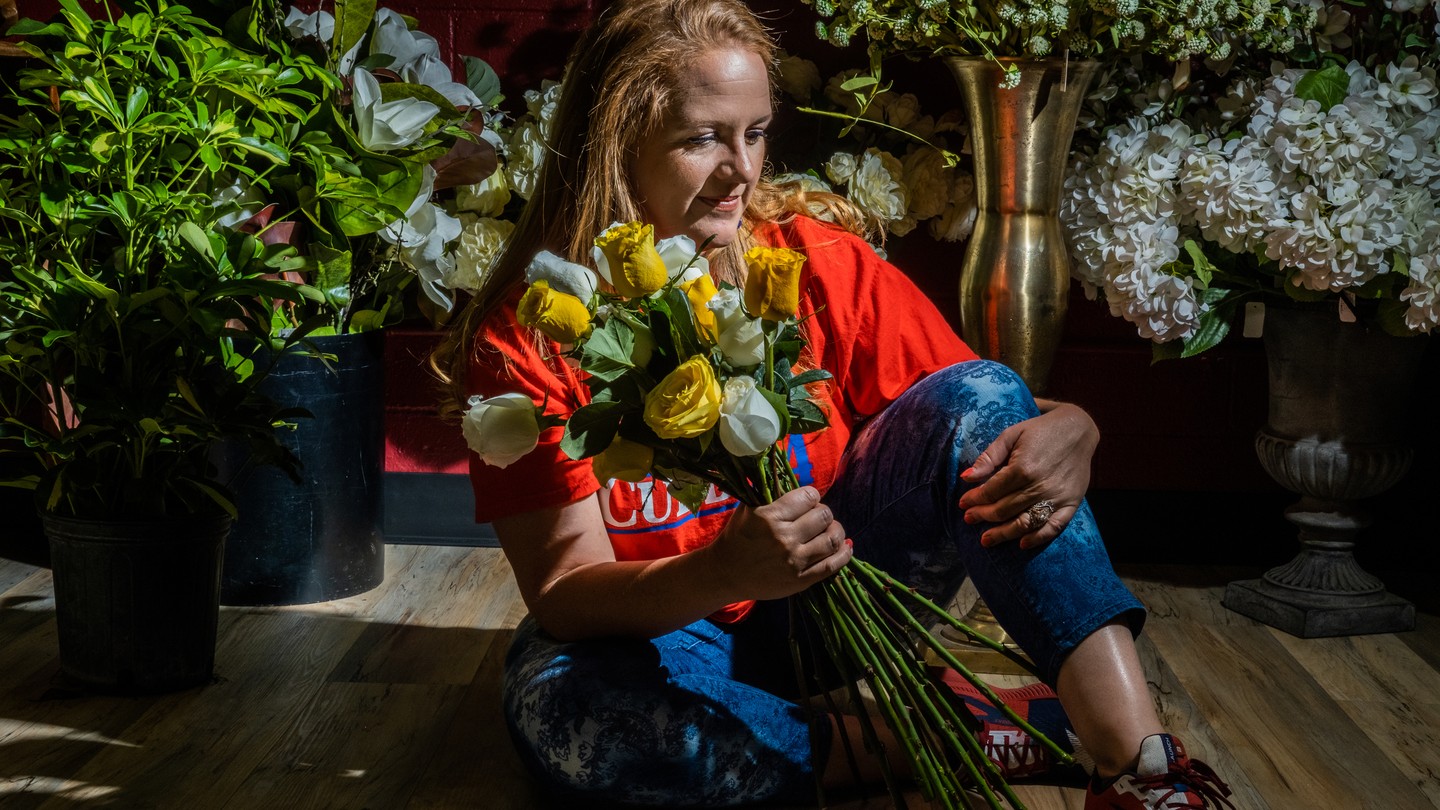 Jenny Cudd sits on the floor of her flower shop in Midland, Texas. She is wearing a red T-shirt and blue pants and holding a bunch of yellow and white long-stemmed roses.